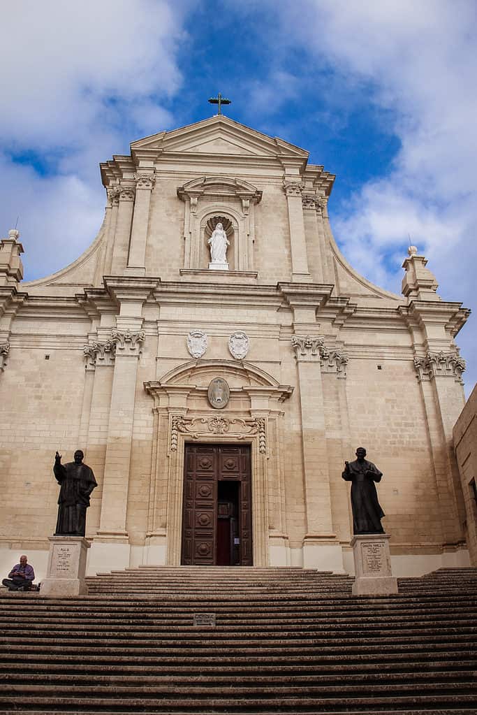 The grand stone façade of a historic Maltese church with tall columns, carved statues, and a wide staircase leading to a large wooden entrance door beneath a cloudy sky.