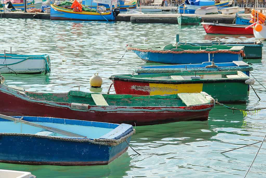Colorful traditional Maltese luzzu boats floating in the calm water of Marsaxlokk Bay, with more boats and a small waterfront area in the background.