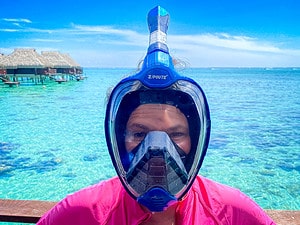 Person wearing a full‑face snorkel mask in front of turquoise water with overwater bungalows in the background.
