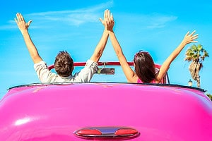 Couple riding in a pink convertible with arms raised, ready for road trip questions for couples.
