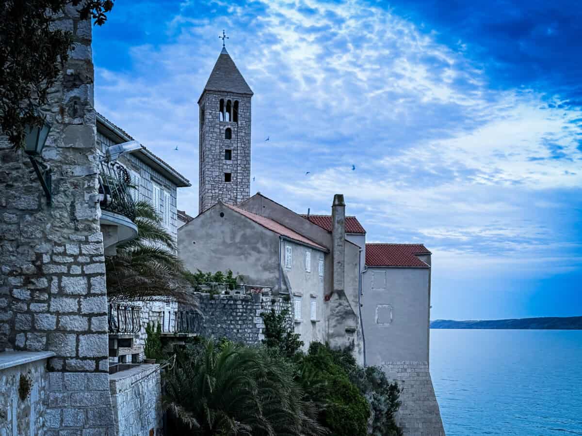 A stone bell tower rises above Rab Old Town's whitewashed buildings, with the Adriatic Sea in the background.