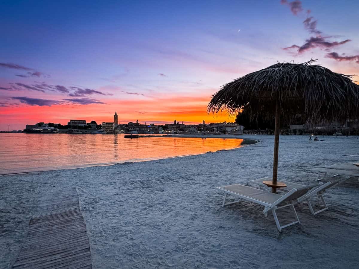 Rab Town's bell tower skyline glows orange and pink at sunset, seen across the water from a nearby beach.