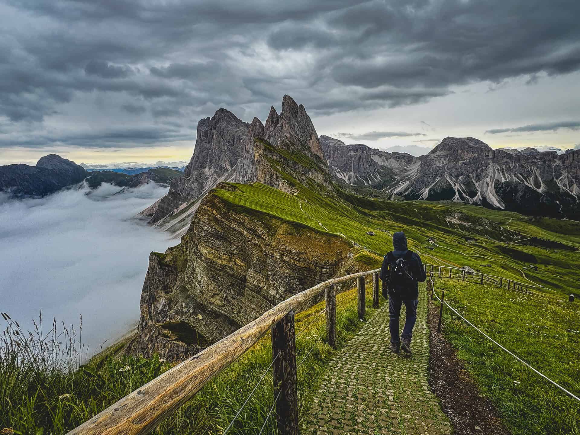 20260821 secedaridge 1869 A hiker walks a cobbled path along the Seceda ridge with the Odle spires and a sea of cloud beyond, in the Dolomites of northern Italy.