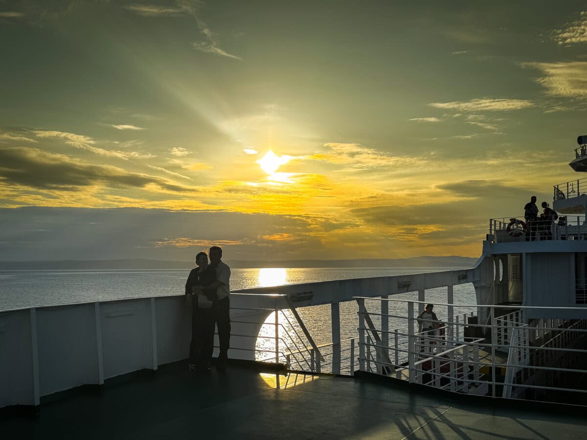A couple watches the sunset from the deck of a car ferry crossing toward Rab Island, the sky glowing gold and orange.