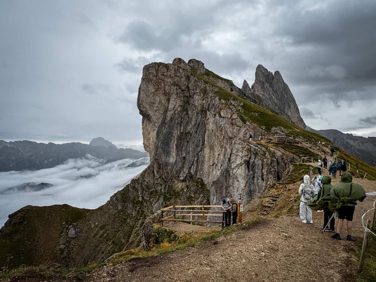 20260821 secedaridge 1879 Hikers in rain gear stand at a Seceda viewpoint with cloud filling the valley below.