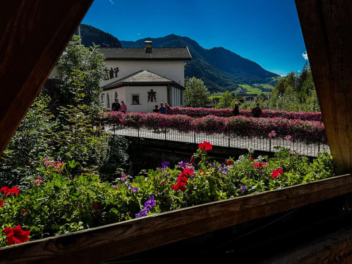 20260822 wheretostayinthedolomites 2193 View of a flower-covered bridge and mountains in Ortisei, Val Gardena, Italy, framed by a wooden balcony.