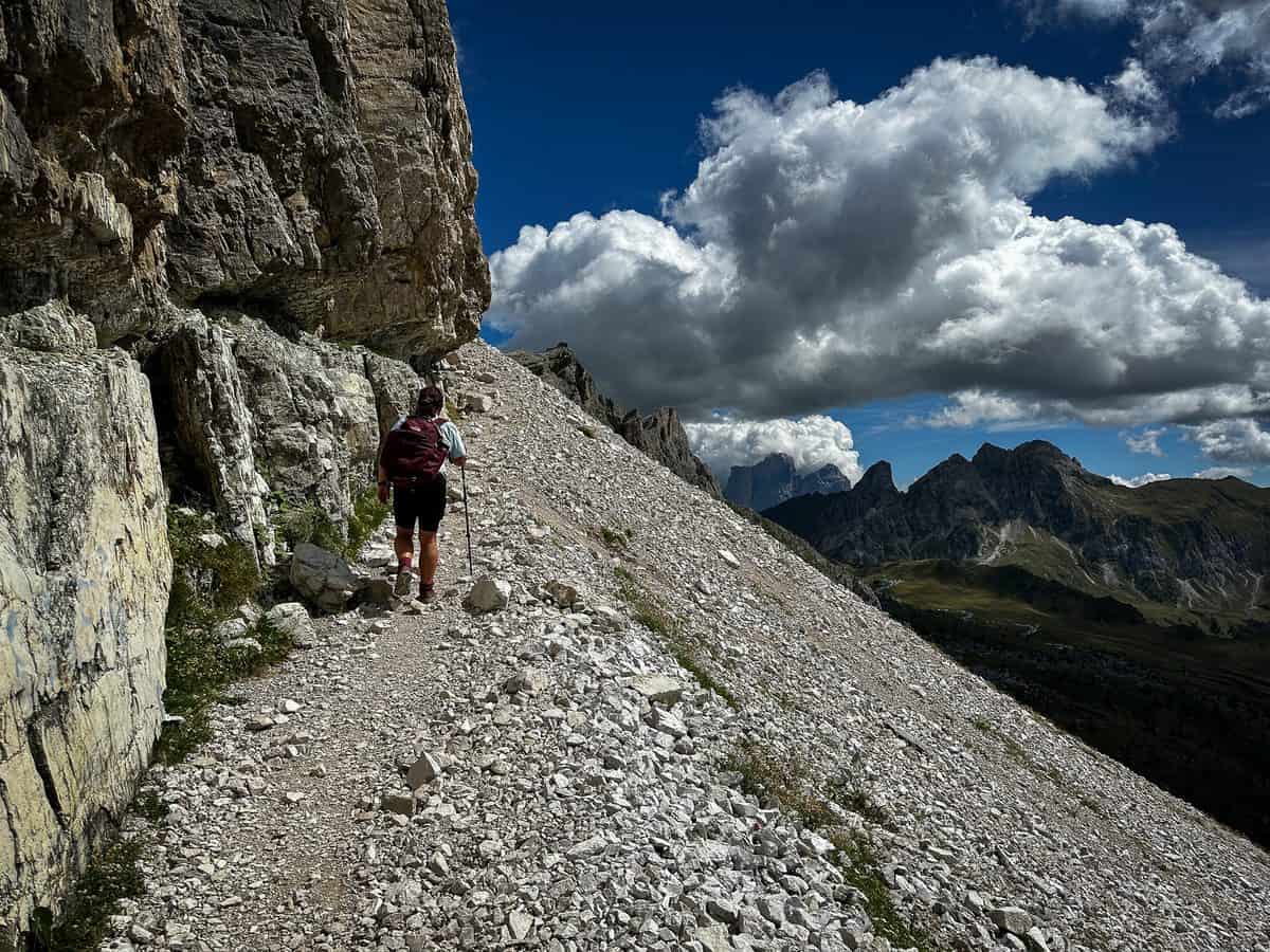 20260826 besthikesinthedolomitesaverau Jolayne crosses a narrow gravel trail cut into the mountainside, with peaks and dramatic clouds in the distance on the Mt. Averau summit trail.