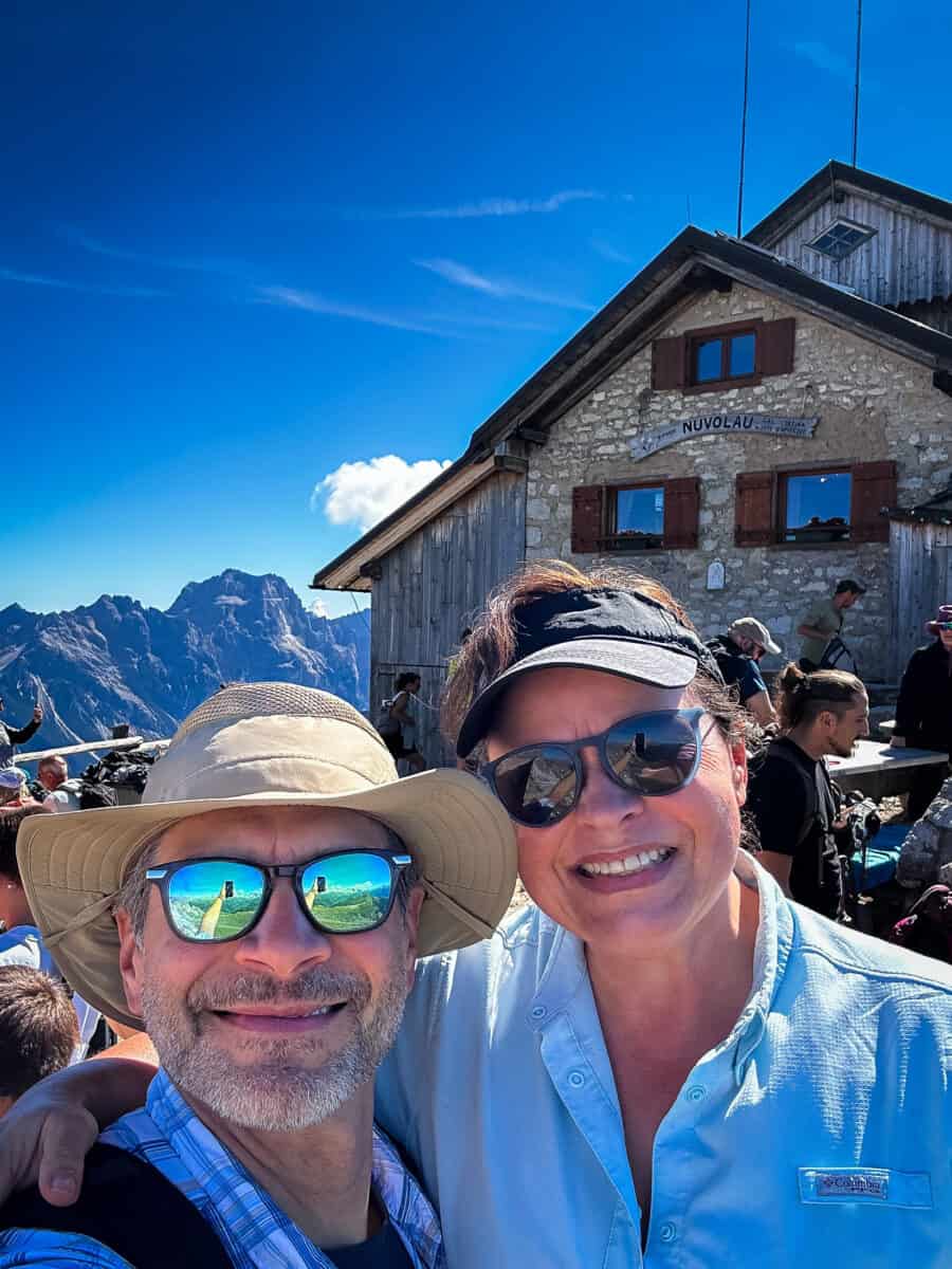 20260826 besthikesinthedolomitesnuvolau Jolayne and Chris smile for a selfie in front of Rifugio Nuvolau, the oldest mountain hut in the Dolomites, with peaks in the background.