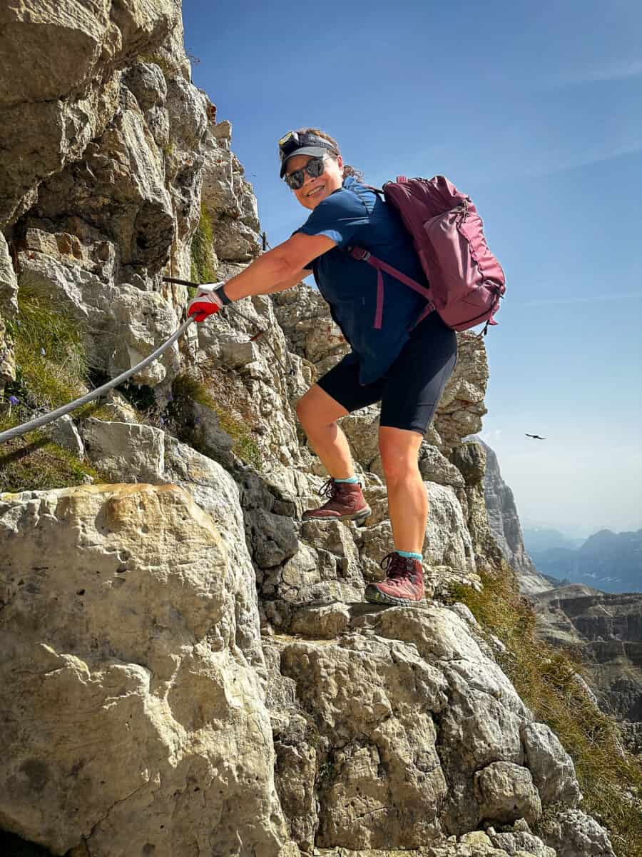 20260827 besthikesinthedolomites 8910 Jolayne climbs a cabled rock section on the Rifugio Lagazuoi tunnel route, gripping the cable with a gloved hand.