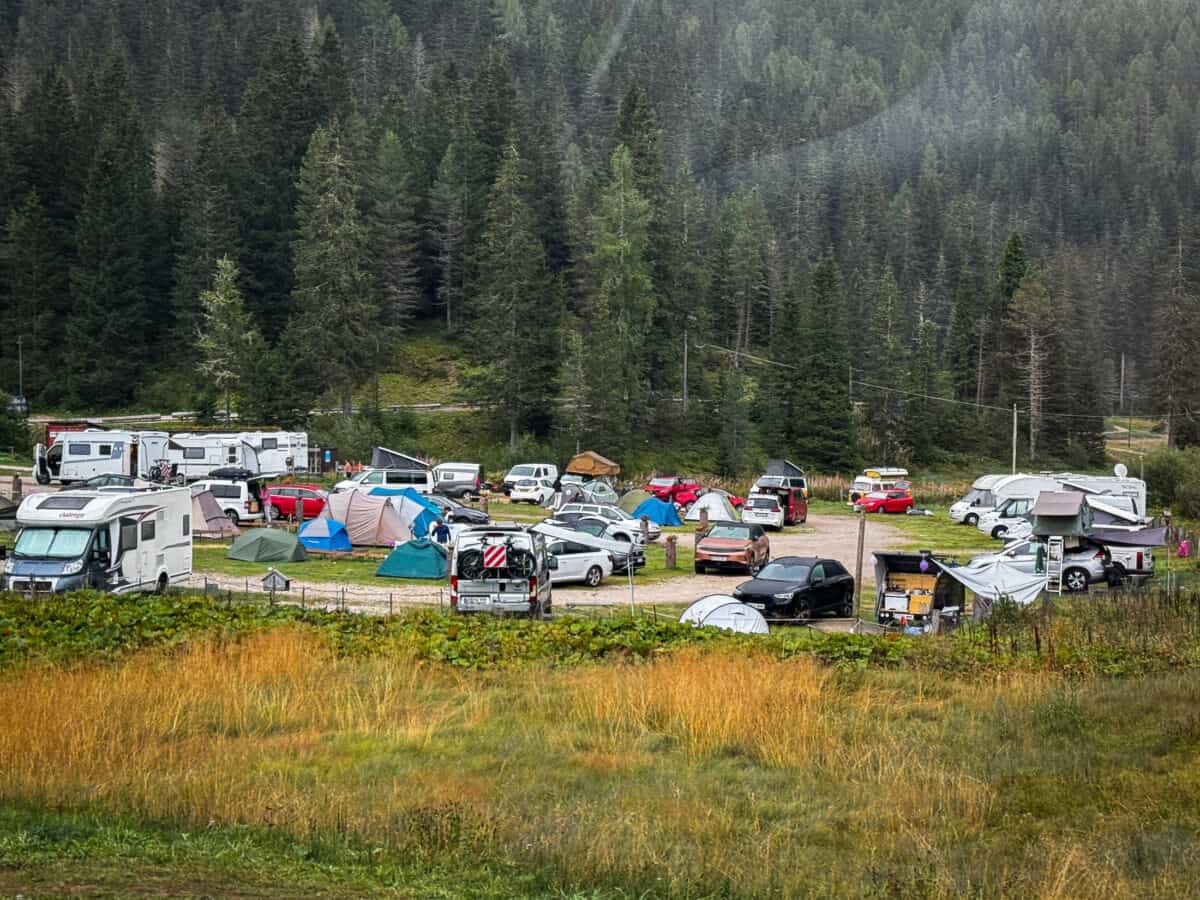 Crowded Camping Alla Baita campground with campervans, tents & cars below a pine forest near Lake Misurina in the Dolomites.