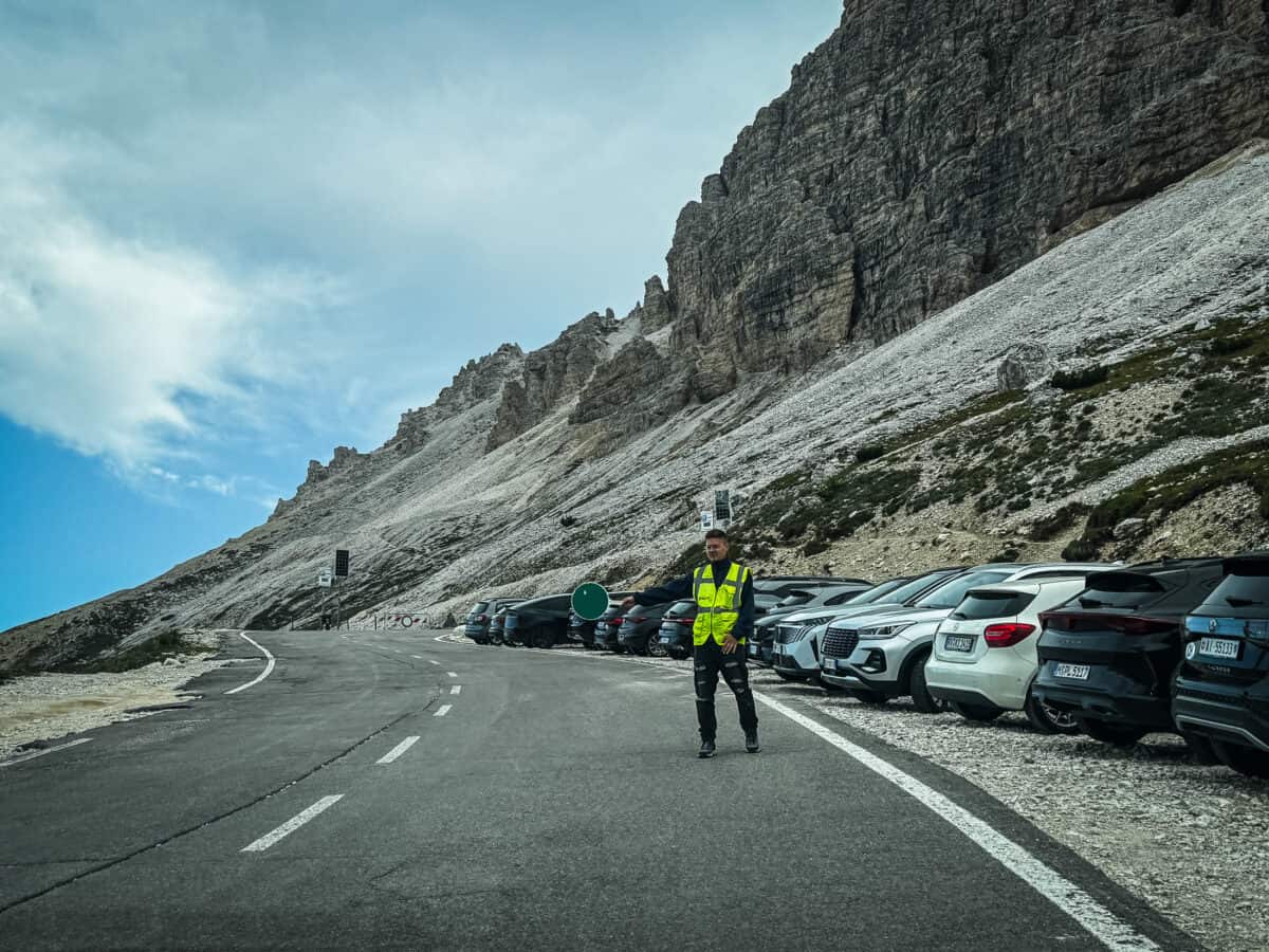 Parking attendant in a yellow vest directing cars to roadside parking below rocky cliffs at the Tre Cime di Lavaredo trailhead.