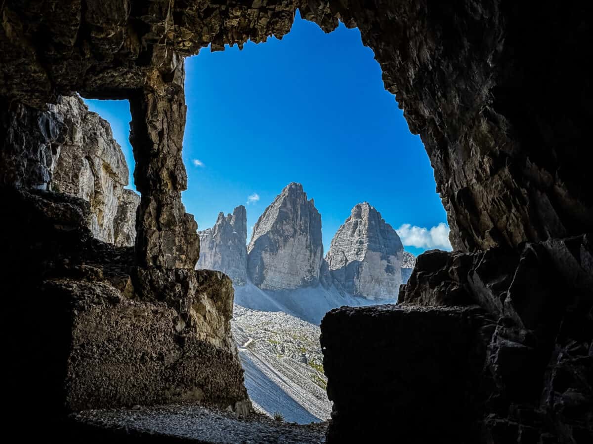 The three peaks of Tre Cime di Lavaredo framed by a rocky tunnel opening in Monte Paterno under a blue sky.