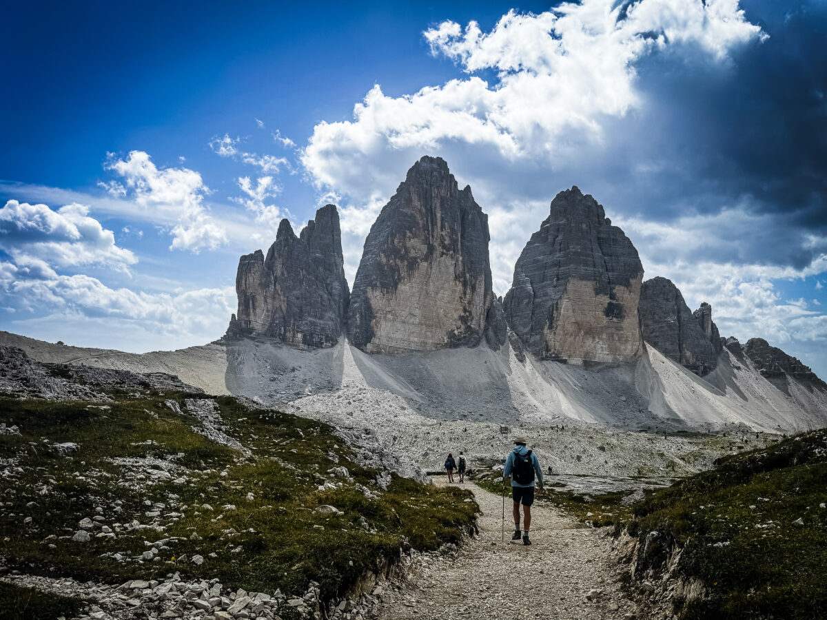 20260828 trecimedilavaredohike 2967 Hiker on the Tre Cime di Lavaredo hike walking toward the three iconic north faces under dramatic clouds in the Dolomites.