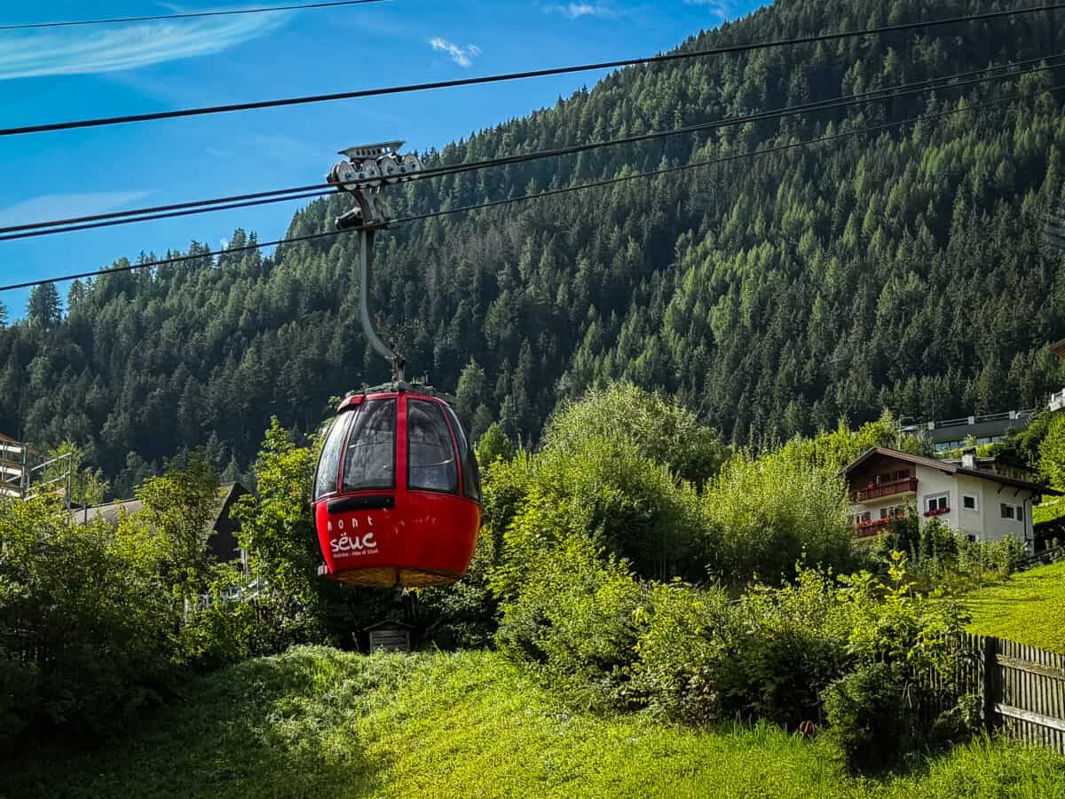 Red Mont Sëuc gondola leaving Ortisei for Alpe di Siusi, above green meadows and village houses.