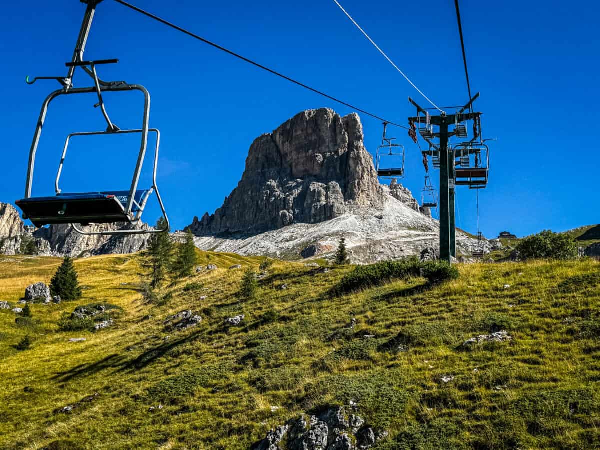 Empty chairs on the Fedare chairlift climbing toward a jagged Dolomites peak under a deep blue sky above Passo Giau.