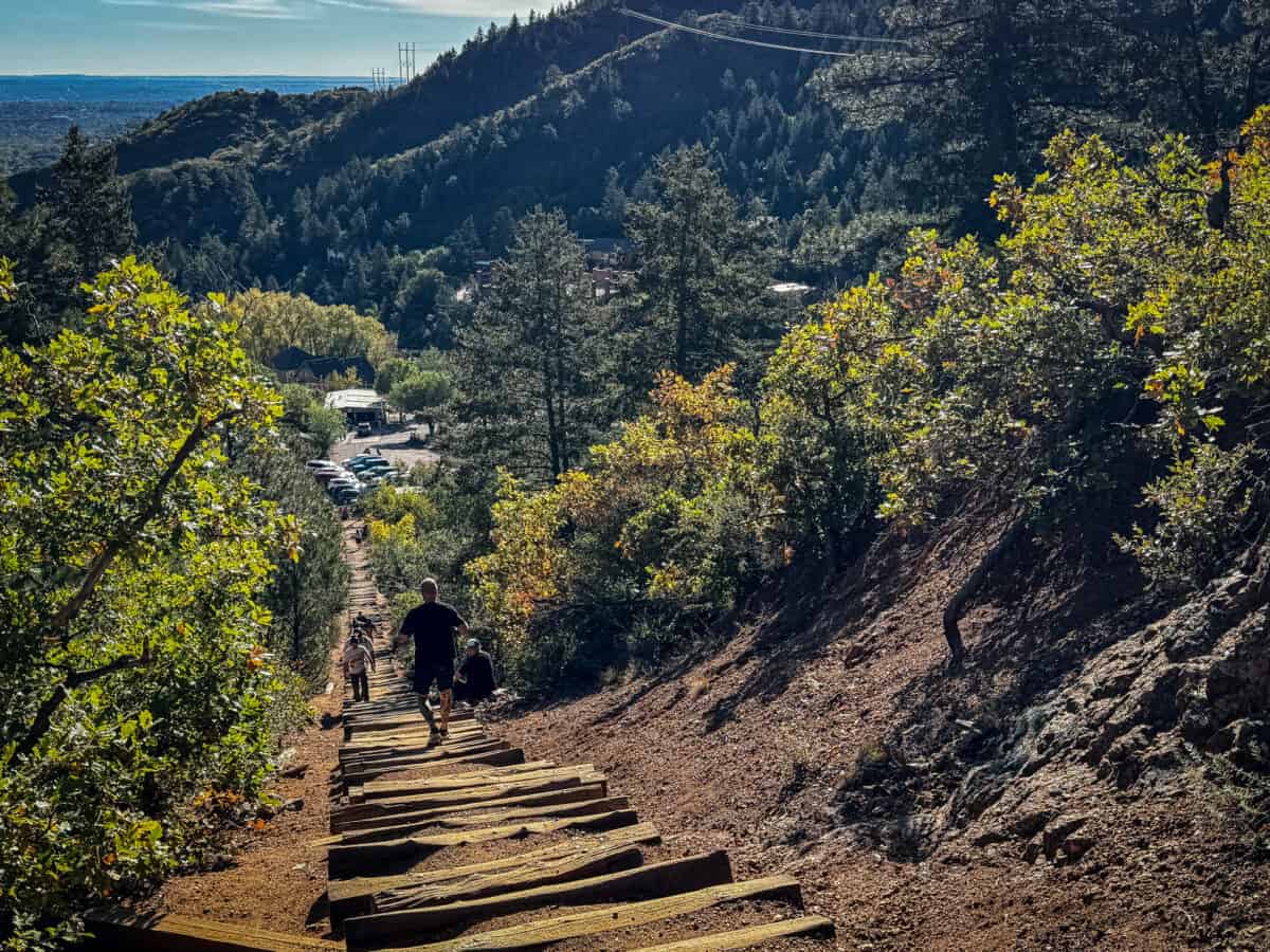 A hiker walking down the Manitou Incline steps toward the trailhead parking lot as other hikers climb up past him through fall foliage.