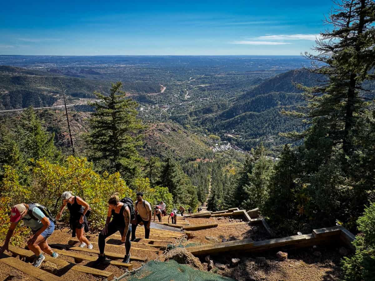 Hikers climbing the railroad-tie steps of the Manitou Incline hike through pine trees under a blue Colorado sky.