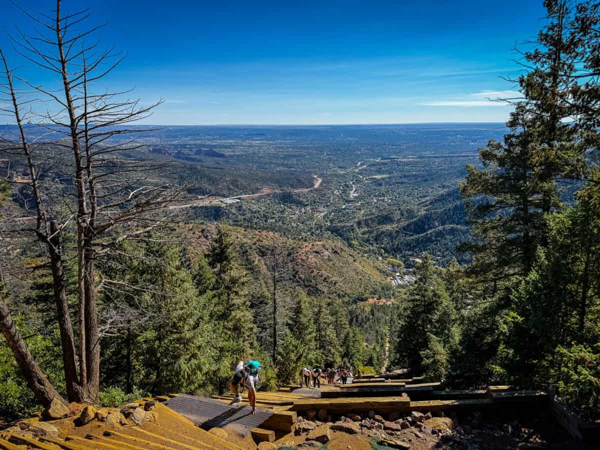 A hiker climbing on hands and feet up a steep section of the Manitou Incline, with Manitou Springs and the plains stretching out far below.