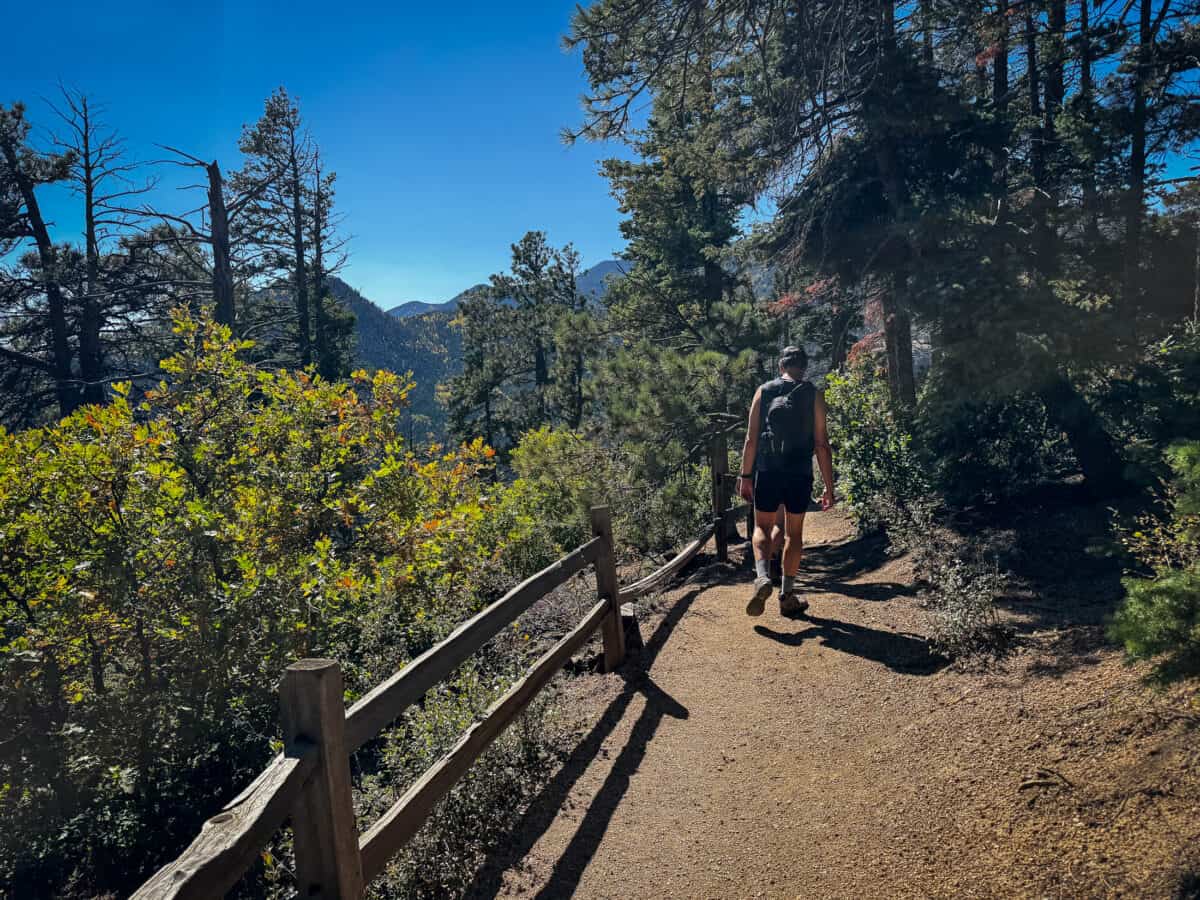 Chris heading down the Barr Trail past a wooden fence, with pine trees and fall foliage under a bright blue sky.